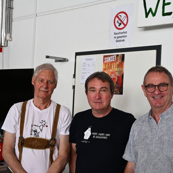 Drei Männer stehen neben einer Wand mit einem Rauchverbotsschild. Der linke Mann trägt ein weißes Shirt mit einer Ziegengrafik, der mittlere Mann trägt ein schwarzes Shirt mit einer weißen Grafik und der rechte Mann trägt ein graues Hemd.