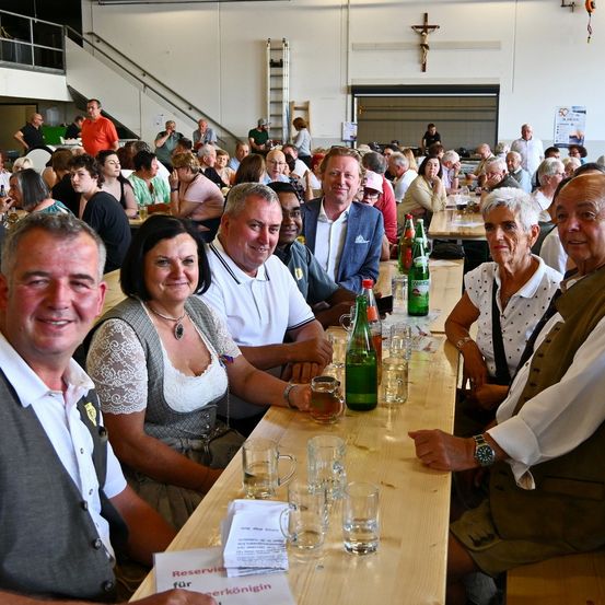 Eine Gruppe von Menschen sitzt an einem langen Tisch in einem Saal, lächelt und schaut zur Kamera. Auf dem Tisch sind Gläser, Flaschen und Papiere. Der Saal hat ein Kreuz an der Wand, eine Treppe und eine Leiter.