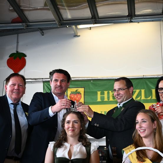 Fünf Männer und eine Frau krönen ein lächelndes Mädchen mit einer Tiara unter einem grünen Banner mit einer Erdbeere darauf.