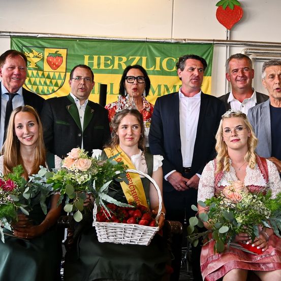 Eine Gruppe von Menschen in traditioneller Kleidung, die Blumensträuße halten und lächeln. Sie stehen vor einem Banner mit Text und einer Erdbeere darauf.