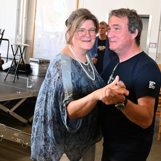 Ein Mann und eine Frau tanzen lächelnd zusammen in einem Innenbereich. Ein Mikrofon wird von einem Mann in einem blauen T-Shirt im Hintergrund gehalten.