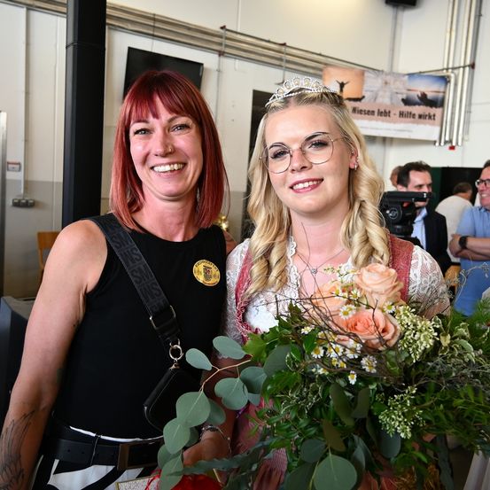 Zwei Frauen stehen lächelnd da, eine mit einem Blumenkranz, halten einen Blumenstrauß. Dahinter hält ein Mann eine Kamera und eine Frau schaut zu. Ein Schild sagt 'Wiesen lebt - Hilfe wirkt.'