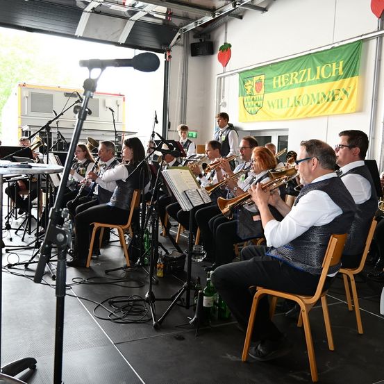 Eine Gruppe von Musikern in einem Raum, die Blechblasinstrumente und andere Blasinstrumente spielen. Ein Banner mit der Aufschrift Herzlich Willkommen ist sichtbar.