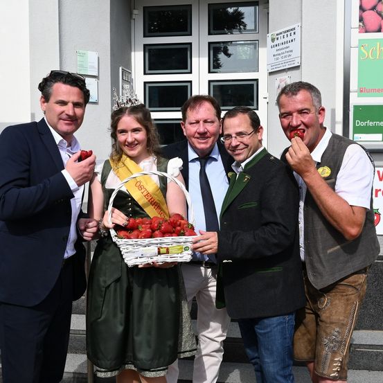 Fünf Personen, darunter eine Frau in traditioneller bayerischer Kleidung, lächeln und posieren für ein Foto, während sie Erdbeeren halten. Sie stehen vor einem weißen Gebäude mit Glasfenstern und Schildern.