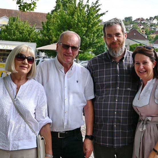 Vier Personen stehen lächelnd draußen für ein Foto. Zwei Männer und zwei Frauen, mit Bäumen und Häusern im Hintergrund.