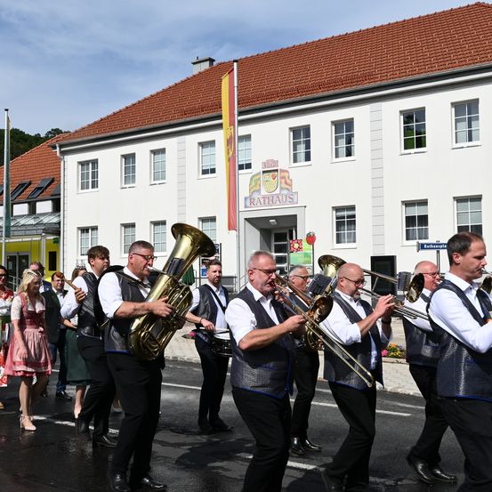 Eine Blechbläsergruppe spielt Musik vor einem großen Gebäude mit rotem Dach. Die Bandmitglieder tragen passende Westen und Hosen. Eine Frau in einem rosa Kleid geht in der Nähe vorbei.