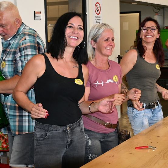 Vier Erwachsene stehen in einem Raum, lächeln und posieren möglicherweise für ein Foto. Drei Frauen stehen vor einem Mann. Die Frauen tragen verschiedenfarbige Shirts und Jeans. Ein rotes Werkzeug liegt auf einem Tisch.