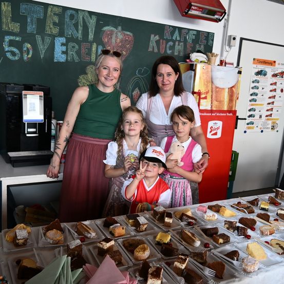 Vier Frauen und zwei junge Mädchen stehen hinter einem Tisch voller Desserts. Die Frau links trägt ein ärmelloses grünes Oberteil und einen kastanienbraunen Rock. Im Hintergrund steht eine Tafel mit der Aufschrift 'Litery 5dverein'.