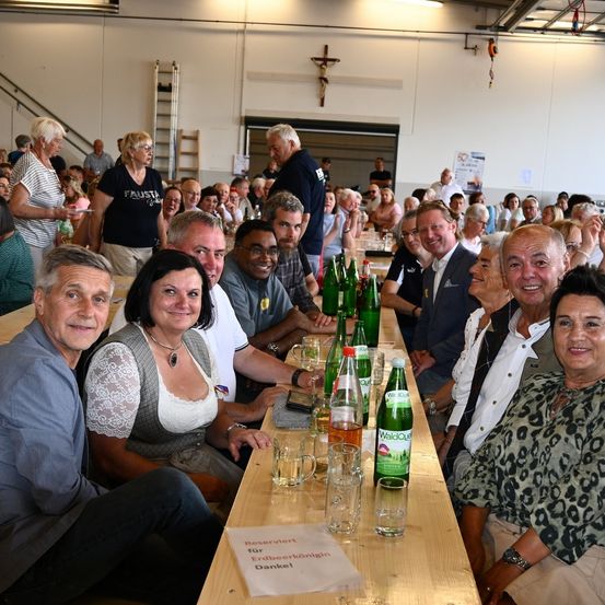 Eine Gruppe von Menschen sitzt an einem langen Tisch in einem Saal, einige lächeln, andere schauen in die Kamera. Flaschen, Gläser und ein Schild, das Reserviert für Erdbeerkönigin Dankel steht, sind auf dem Tisch. Ein Kreuz hängt an der Wand hinter ihnen.