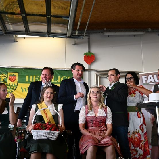 Eine Gruppe von Menschen, darunter zwei Frauen in traditioneller bayerischer Kleidung, steht in einem Raum mit einem Erdbeerbanner. Eine Frau hält einen Korb mit Erdbeeren. Ein Mann justiert eine Krone auf dem Kopf einer Frau. Eine andere Frau justiert die Brille eines Mannes.