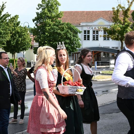 Drei Frauen in traditioneller bayerischer Kleidung gehen auf der Straße. Eine Frau trägt eine Krone, eine andere hält einen Korb mit Erdbeeren. Hinter ihnen stehen mehrere Menschen, darunter ein Mann mit Rucksack.