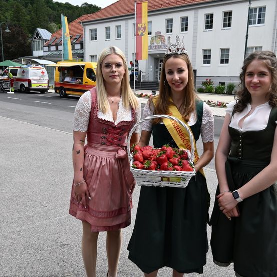 Drei Frauen in traditioneller Kleidung stehen auf einer Straße, eine hält einen Korb mit Erdbeeren. Hinter ihnen steht ein weißes Gebäude mit Fahnen, und ein gelber und weißer Van ist geparkt.
