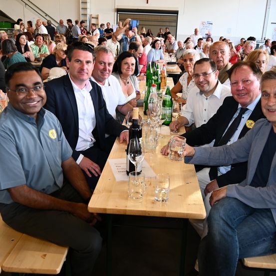 Eine Gruppe von Menschen sitzt an einem Tisch, wahrscheinlich bei einer Veranstaltung, mit mehreren Weinflaschen, Gläsern und einem Namensschild. Einige lächeln, während andere Fotos machen.