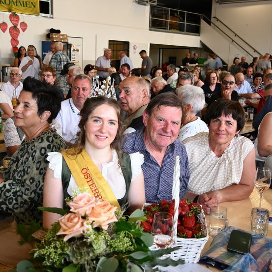 Eine Gruppe von Menschen ist bei einer Veranstaltung versammelt, sitzt an einem Tisch mit Blumen, Erdbeeren und Getränken. Eine Frau in einem Band sitzt in der Mitte.