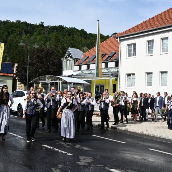 Eine Blaskapelle, möglicherweise für eine Parade, geht auf der Straße. Sie wird von Menschen in traditioneller Kleidung und formeller Kleidung begleitet. Im Hintergrund befinden sich Gebäude, Bäume und ein Fahnenmast.