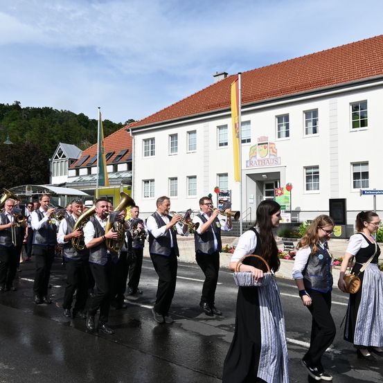 Eine Blaskapelle spielt vor einem großen weißen Gebäude mit rotem Dach. Das Gebäude trägt ein Schild mit der Aufschrift Rathaus. Die Bandmitglieder tragen traditionelle deutsche Kleidung.