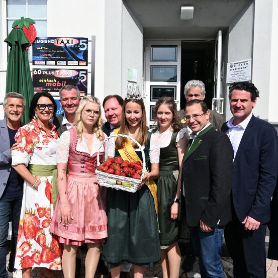 Eine Gruppe Erwachsener in traditioneller Kleidung steht vor einem Gebäude. Eine Frau hält einen Korb mit Erdbeeren. Ein Plakat, das Jugendtaxi und 60 Plus Taxi bewirbt, ist sichtbar.