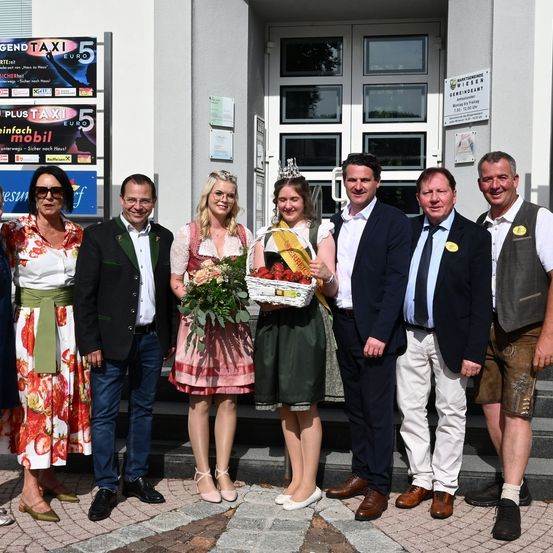 Eine Gruppe Erwachsener, darunter zwei Frauen in traditionellen deutschen Kleidern, steht vor einem Gebäude. Sie lächeln alle und posieren für ein Foto. Die Frau in der Mitte hält einen Korb mit Blumen und eine Krone.