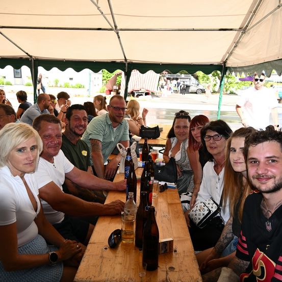Eine Gruppe von Menschen versammelt sich unter einem Zelt bei einer Outdoor-Veranstaltung. Sie sitzen um einen Tisch herum, der mit Flaschen und Gläsern gefüllt ist. Einige tragen Sonnenbrillen, und eine Person macht ein Foto. Im Hintergrund gehen Menschen auf einer nassen Straße.