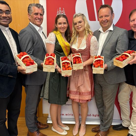 Eine Gruppe von Menschen in formeller Kleidung hält Körbe mit Erdbeeren vor einem roten Hintergrund mit Logo. Zwei Frauen tragen Kronen.