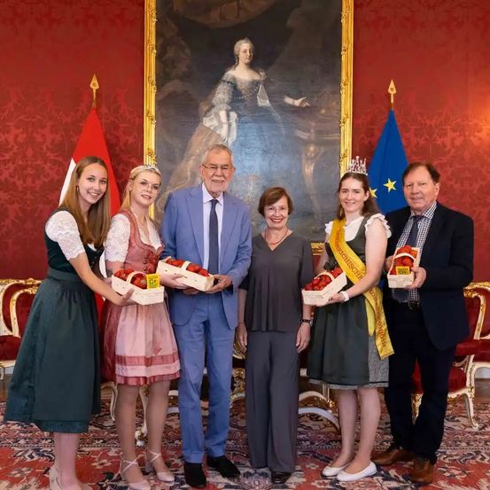 Eine Gruppe von Menschen, darunter zwei Frauen in traditionellen Kleidern, steht vor einem großen Porträt einer Frau. Sie halten Körbe mit Erdbeeren.
