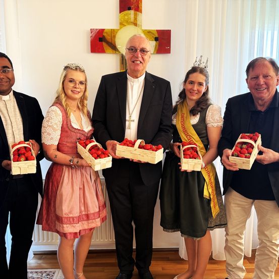 Fünf Personen, darunter zwei Frauen in traditionellen Kleidern, stehen zusammen und halten Erdbeerkörbe, wobei ein Priester in der Mitte eine Halskette und ein Kreuz trägt. Hinter ihnen ist ein Kreuz an der Wand befestigt.