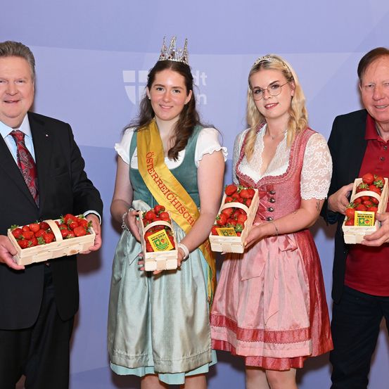Vier Personen, zwei Männer und zwei Frauen, stehen zusammen und halten Körbe mit Erdbeeren. Die Frauen tragen traditionelle bayerische Kleider und Schärpen, eine mit einer Krone. Die Männer tragen Anzüge und Krawatten.