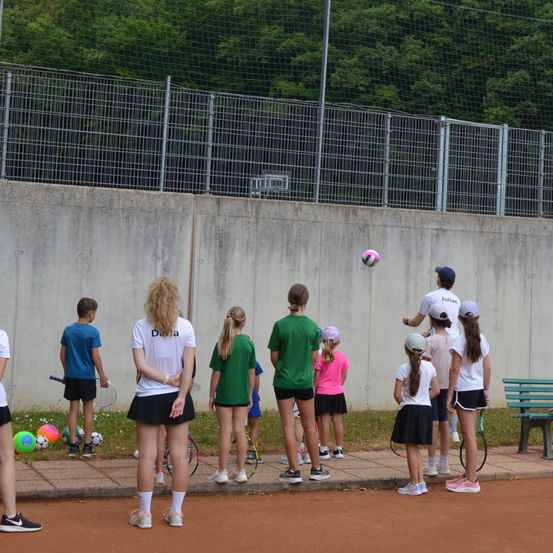 Eine Gruppe junger Menschen steht auf einem Tennisplatz und beobachtet, wie ein Ball in die Luft geworfen wird. Einige von ihnen halten Tennisschläger.