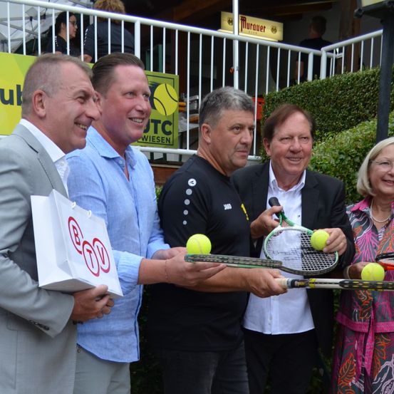 Fünf Personen stehen zusammen und halten Tennisschläger und Tennisbälle. Ein Mann im Anzug hält eine Tasche mit einem roten Logo. Hinter ihnen steht ein Schild mit den Buchstaben 'WTC'.