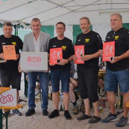 Fünf Männer stehen unter einem Zelt, halten Bücher und Pokale. Sie sind in schwarzen T-Shirts und Shorts gekleidet. Der Mann in der Mitte hält eine Box mit der Aufschrift BTV.