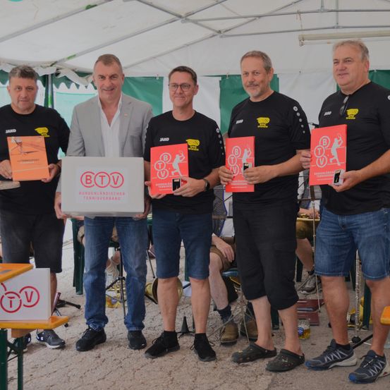 Fünf Männer stehen unter einem Zelt, halten Bücher und Pokale. Sie sind in schwarzen T-Shirts und Shorts gekleidet. Der Mann in der Mitte hält eine Box mit der Aufschrift BTV.