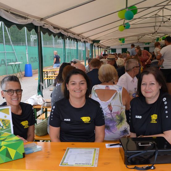 Drei Frauen in schwarzen T-Shirts mit gelben Logos posieren für ein Foto in einem Zelt mit einer Menschenmenge im Hintergrund.