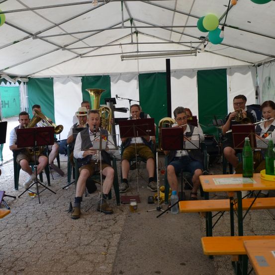 Eine Gruppe von Musikern in traditioneller Kleidung spielt in einem Zelt, mit verschiedenen Instrumenten wie Tubas und Trompeten. Die Umgebung scheint ein Festival oder eine Veranstaltung im Freien zu sein.