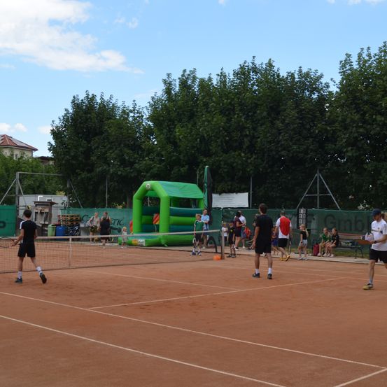 Eine Gruppe von Menschen spielt Tennis auf einem Sandplatz. Einige spielen, während andere zuschauen. Im Hintergrund befindet sich ein grünes aufblasbares Hindernis.