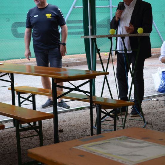 Ein Mann in einem schwarzen T-Shirt mit einem gelben Logo steht hinter einem Tisch mit Tennisbällen darauf, während ein anderer Mann in einem Anzug in ein Mikrofon spricht.