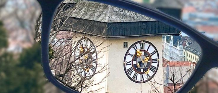 Eine Brille zeigt eine Reflexion eines Uhrturms. Die Uhr hat römische Zahlen. Bäume umgeben den Turm. Der Turm befindet sich in der Mitte einer Stadt.