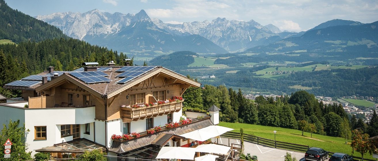 Ein Chalet mit Solarpanels auf dem Dach steht auf einem Hügel, umgeben von Bergen, üppigen grünen Feldern und einem klaren blauen Himmel.