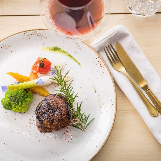 Ein Teller mit Steak, garniert mit Rosmarin und Gemüse, begleitet von einem Glas Wein auf einem Holztisch.