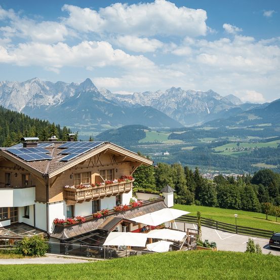 Ein Chalet mit Solarmodulen auf dem Dach bietet Blick auf ein Tal und Berge. Es verfügt über einen Balkon mit Blumenkästen und einen Außenbereich mit Sitzgelegenheiten und Sonnenschirmen.