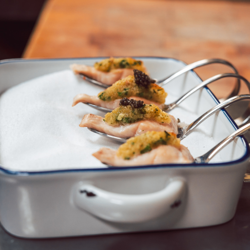 Fünf Stück Fisch, wahrscheinlich Forellen, sind auf Löffeln in einer weißen Schale, überzogen mit grüner Beilage und einer Scheibe schwarzen Kaviars. Die Schale ist mit Zucker gefüllt.