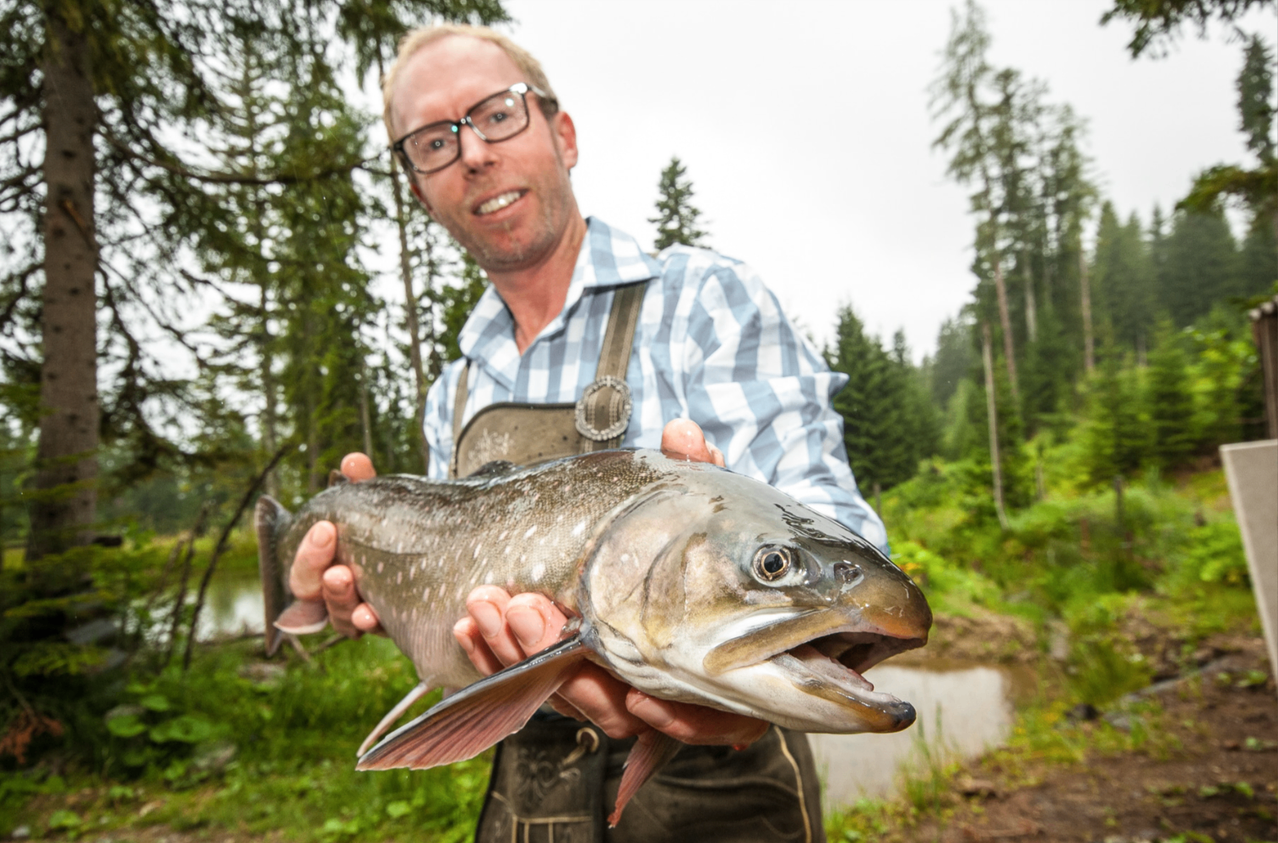 Ein Mann hält einen großen Fisch in einem Wald in der Nähe eines Teichs. Er trägt ein blau kariertes Hemd und eine Brille.