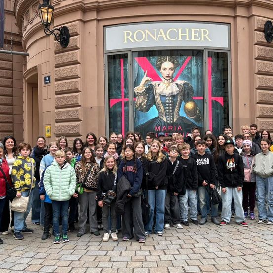 Eine Gruppe von Schülern posiert vor einem Geschäft mit einem großen Fenster, das ein Porträt von Königin Maria zeigt.