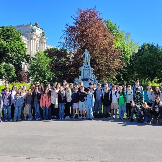 Eine Gruppe von Schülern posiert für ein Foto vor einer Statue in einem Park. Die Statue ist von Bäumen und einem großen Gebäude umgeben.