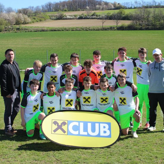 Eine Jugendfußballmannschaft posiert für ein Foto auf einem grünen Feld mit einem Banner, das X Club sagt. Die Mannschaft trägt einheitliche Trikots mit gelben und grünen Akzenten.