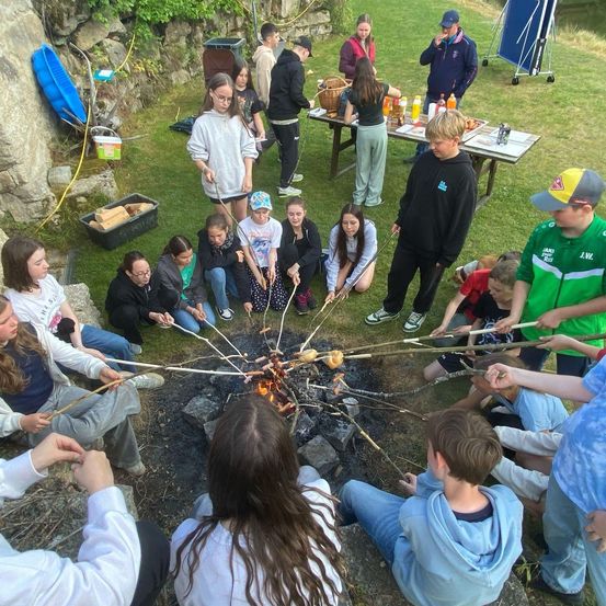 Eine Gruppe von Kindern und Erwachsenen versammelt sich um ein Lagerfeuer und röstet Marshmallows an Stöcken. Einige sitzen auf dem Boden, während andere in der Nähe stehen. Im Hintergrund befinden sich Tische mit Essen und Getränken.