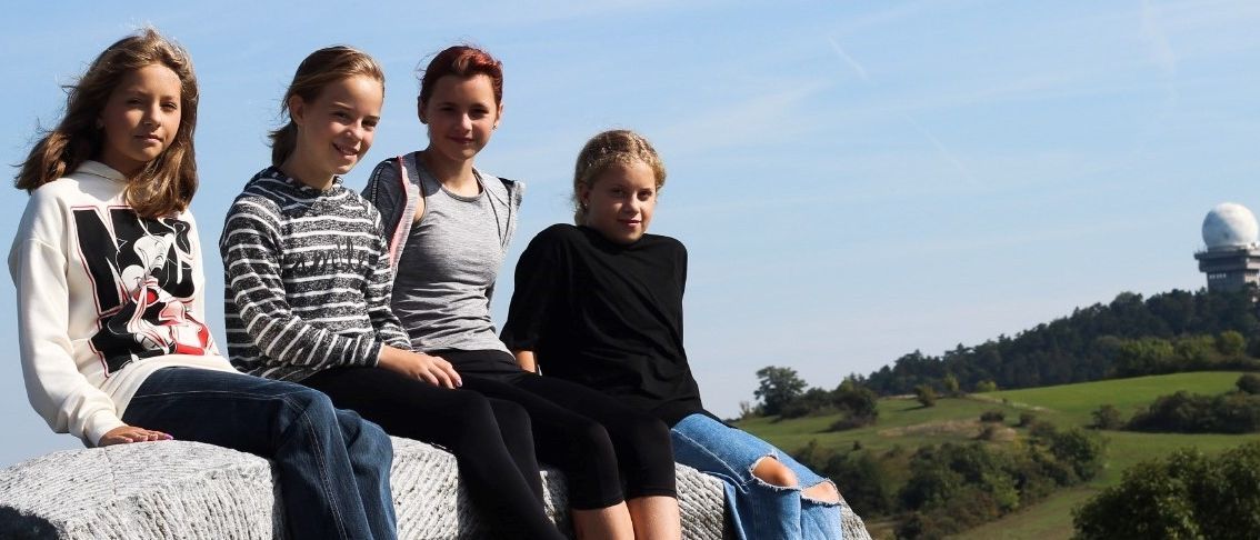 Zwei Mädchen sitzen auf einer weißen Struktur mit Blick auf ein großes kuppelförmiges Gebäude im Hintergrund.