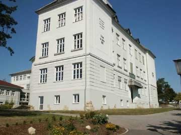 Ein großes weißes Gebäude mit vielen Fenstern, möglicherweise eine Schule, umgeben von Bäumen, Gras und einer gepflasterten Straße.