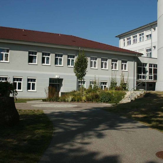 Ein graues Gebäude mit rotem Dach und mehreren Fenstern ist zu sehen. Ein Baum steht davor und ein Betonweg führt zum Eingang hinauf.