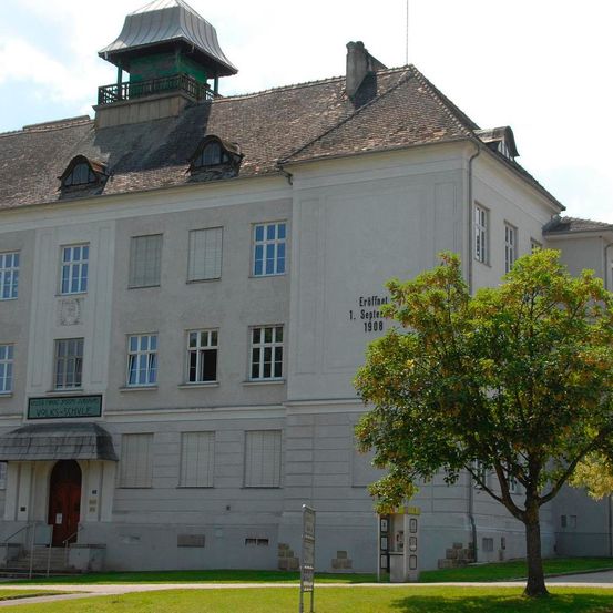 Ein historisches Gebäude mit einem grünen Turm und einem Schild mit der Aufschrift 'Eroffnet 1. September 1908'. Es hat viele Fenster, eine Tür mit einem Vordach und einen Rasen.