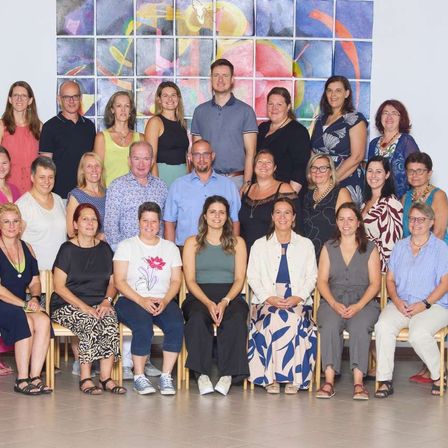 Eine Gruppe von Erwachsenen, sowohl Männer als auch Frauen, sitzen auf Stühlen und stehen in einem Raum, wahrscheinlich für ein Gruppenfoto posieren. Ein farbenfrohes Wandbild befindet sich hinter ihnen.